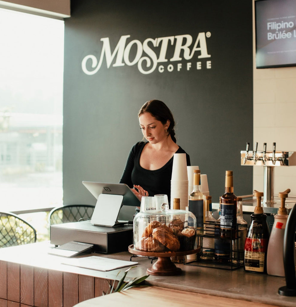 Woman working behind a counter with 'Mostra Coffee' branding in the background.